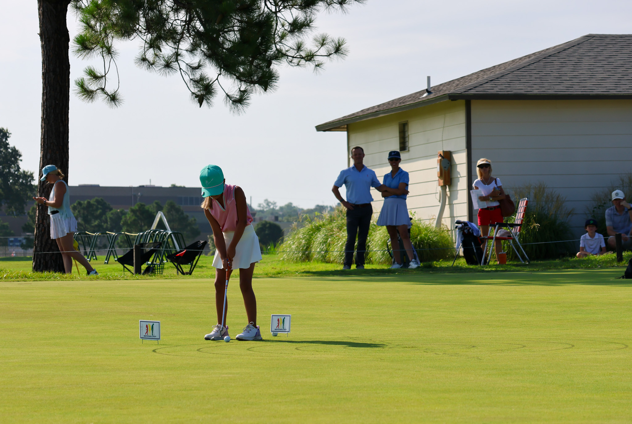 A young girl in a teal cap putts on a sunlit golf course, watched by spectators near a small building. The scene conveys focus and calm.